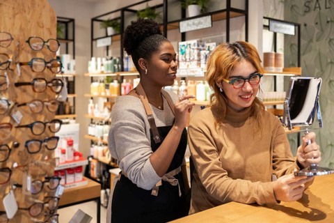 Two women in a cosmetics store engage in a friendly interaction. One is holding a mirror, while the