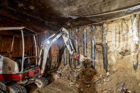 An excavator operates in an underground construction site, tearing through rock and earth. The dim