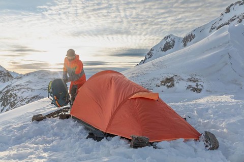 A ski tourer sets up near a bright orange tent amidst snowy mountains at sunrise, capturing the
