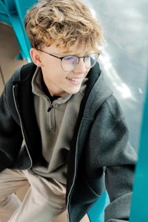 A curly-haired teenager with glasses sits by a window, smiling as he enjoys a ski resort in the