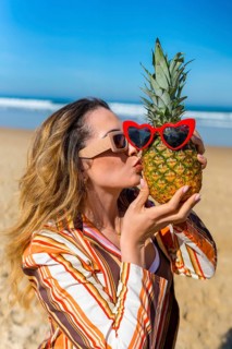 Woman playing with a pineapple wearing red heart sunglasses, standing on a sandy beach by the