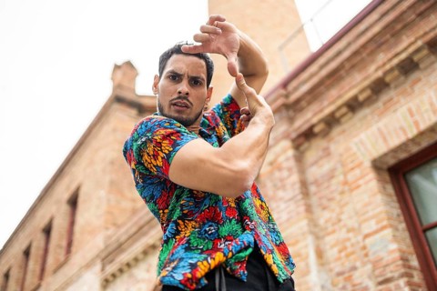 A male dancer in a vibrant, patterned shirt strikes a dynamic pose against a brick building