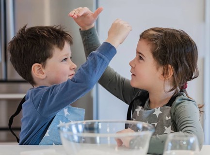 Two young siblings wearing star patterned aprons are joyfully interacting in a kitchen setting. A