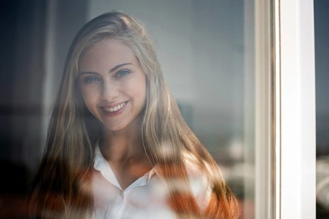 A young woman smiles gently through a glass window, captured in soft natural light. Her relaxed