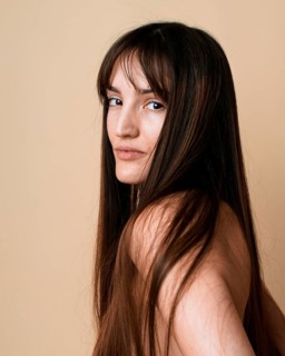 A portrait of a young woman with long brown hair, looking at camera over her shoulder on a beige