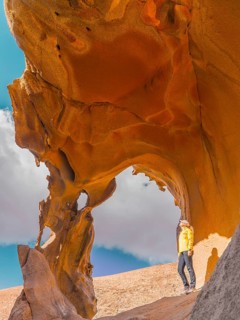 A woman in a bright yellow jacket explores a striking natural rock arch in a desert landscape of