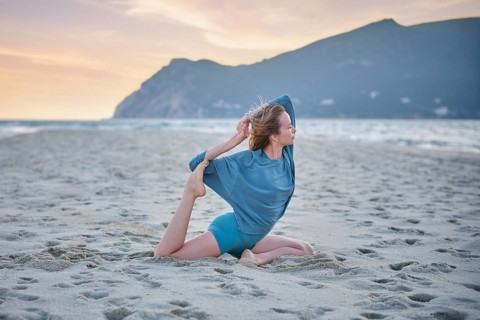 A woman does Eka Pada Rajakapotasana on a sandy beach while the sun sets over the water and hills