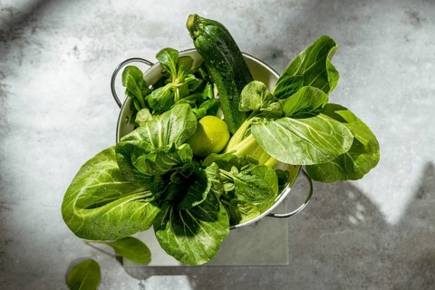 A fresh zucchini, lime, spinach and bok choy in a colander on a gray surface. The vegetables are