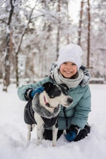 A cheerful child in winter clothing kneels in the snow with a friendly dog. Snow-covered trees