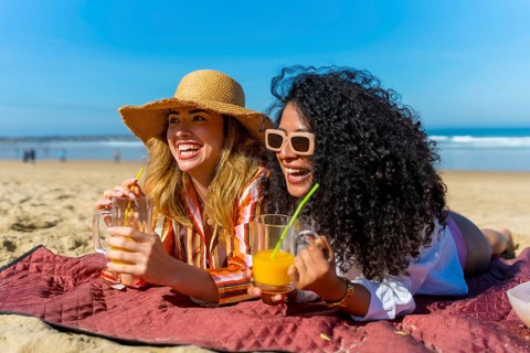 Two cheerful young women lying on a red blanket at the beach, laughing and enjoying their drinks