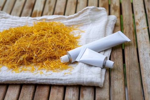 Marigold petals on a cloth and white cosmetic tubes of marigold petals cream on a wooden surface.