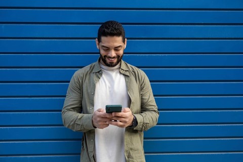 A man in a green shirt and white T shirt smiles while using his smartphone in the city. He stands