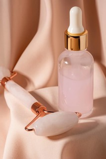A sleek jade roller and a glass dropper bottle containing pink serum sit on elegant fabric These