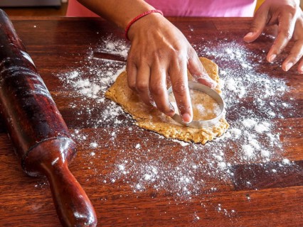 Hands using a circular cutter to shape cookie dough on a floured wooden surface. A rolling pin is
