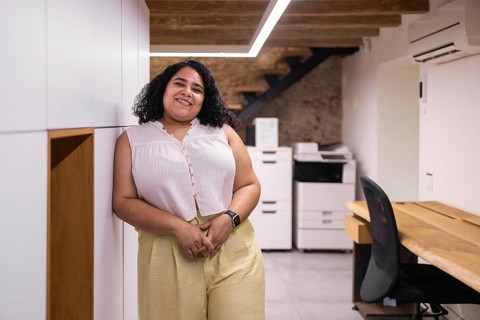 A young woman stands in a modern coworking space with a warm smile. The open office setting is