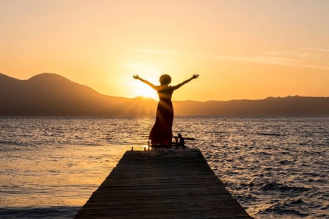 A woman stands on a wooden pier at Papikinou Beach, Milos Island, Greece, embracing the sun as it