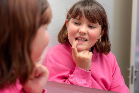 A young girl in a pink sweater smiles as she inspects a loose milk tooth, using a mirror to get a