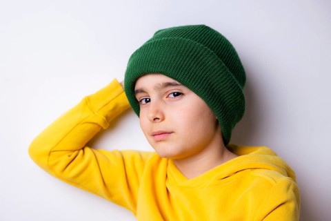 A young child wearing a green knit beanie and a bright yellow hoodie leans against a white wall.