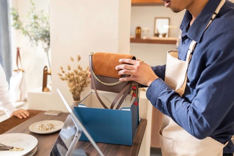 A retail assistant carefully places a stylish handbag into a blue box at a boutique bag store,