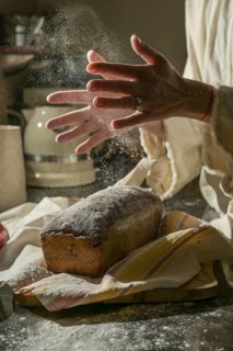 A warm, rustic scene of baking bread. Hands clap to dust flour while a loaf of bread cools on a
