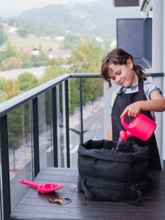 A young girl is carefully watering a sunflower in a black grow bag on a balcony. She is smiling