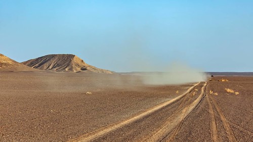 A dusty track runs through a wide, flat desert landscape with hills in the background, with an