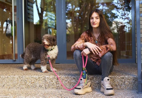 A young woman sits on steps next to a dog with a pink leash, enjoying a sunny day. The scene