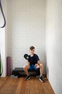 A young boy sitting on an exercise bench lifts a dumbbell in a home gym setting. He is wearing