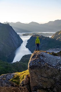 Mountaineer, young man standing on rocks and looking into the distance, at sunrise, fjord