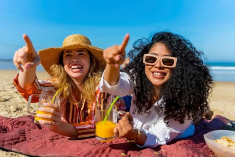 Two cheerful young women lying on a beach blanket, pointing into the distance with excitement while