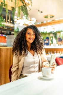 Vertical photo of a happy latin woman using phone while drinking coffee in a modern cafeteria