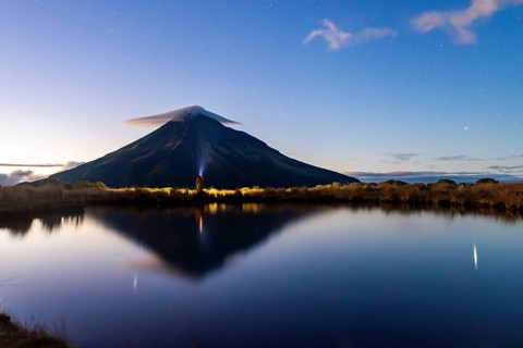 Back view of unrecognizable person looking at Mount Taranaki's reflection in a tranquil lake. The