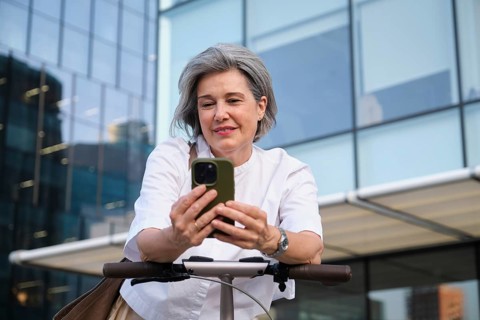 A mature woman with gray hair smiles as she uses a smartphone while standing with an electric