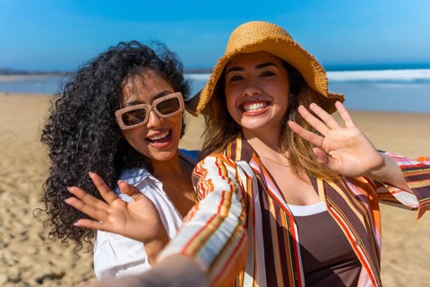 Two cheerful young women on a sunny beach vacation take a joyful selfie, smiling and waving at the