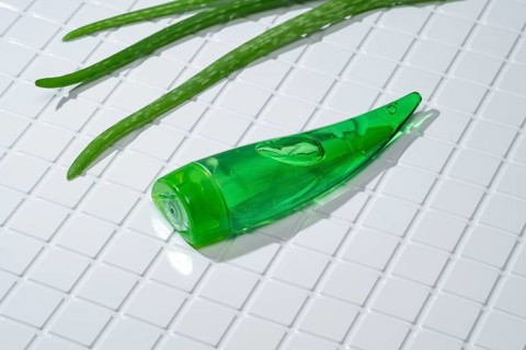 A green aloe vera gel bottle lies alongside fresh aloe leaves on a clean, white-tiled bathroom