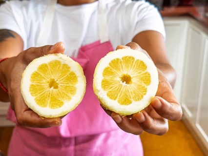 A woman in a pink apron presents two fresh lemon halves in a kitchen setting. The vivid yellow