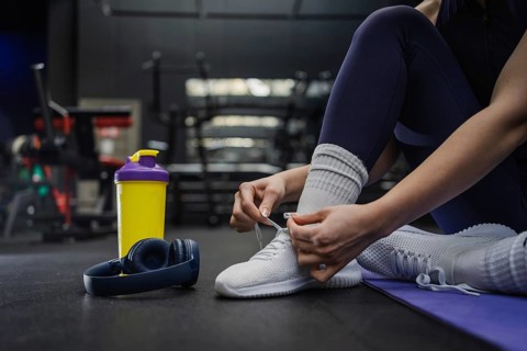 A woman ties their white sneakers, preparing for a workout session. Nearby are wireless headphones