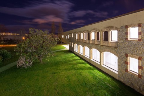 Night view of a well lit hospital with stone and brick facade. The scene includes a green lawn, a