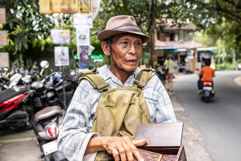 An elderly balinese man wearing a hat and plaid shirt stands on a street, holding a wooden box.
