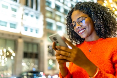 Young woman with glasses and curly hair smiles while using her smartphone on a city street at