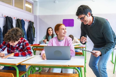 Smiling teacher assisting a student using a laptop in a classroom, other students studying and