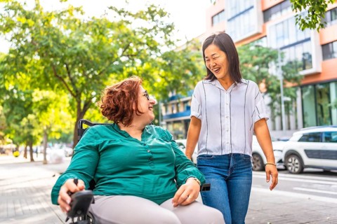 Caucasian happy woman with disability using wheelchair talking while walking with an asian friend