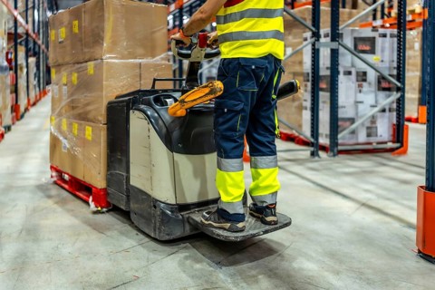 Part of an unrecognizable worker wearing reflective clothes using forklift in a warehouse to move