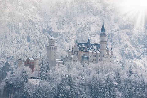 Impressive castle surrounded by snow-covered trees and mountains, with light-flooded sky,