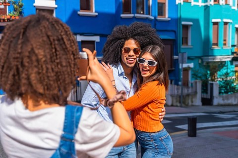 Young woman taking pictures of her two best friends embracing, visiting a colorful city in a sunny