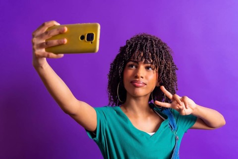 Cheerful young woman with curly hair taking selfie with smartphone and showing victory sign against