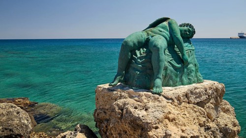 Bronze sculpture, sleeping Eros, sculpture on a rock by the sea with blue water, harbour area,