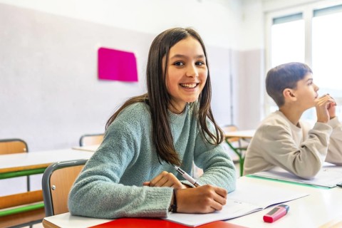 Happy schoolgirl writing on notebook during lesson in classroom with schoolmate thinking in