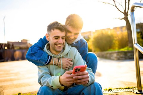 Two men embracing and laughing together while enjoying a smartphone outdoors on a sunny day,