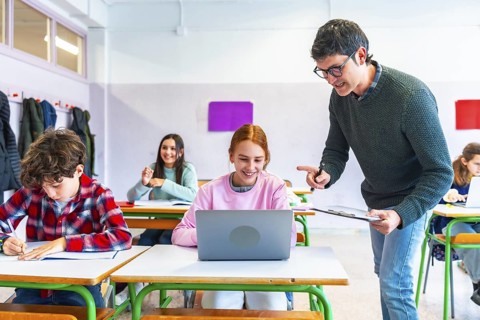Teacher helping a smiling student using laptop computer in classroom during lesson, education and